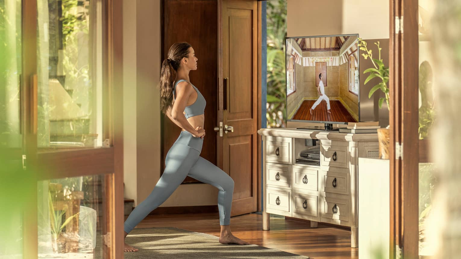 A person in activewear follows a virtual yoga class on a TV screen in a sunlit room