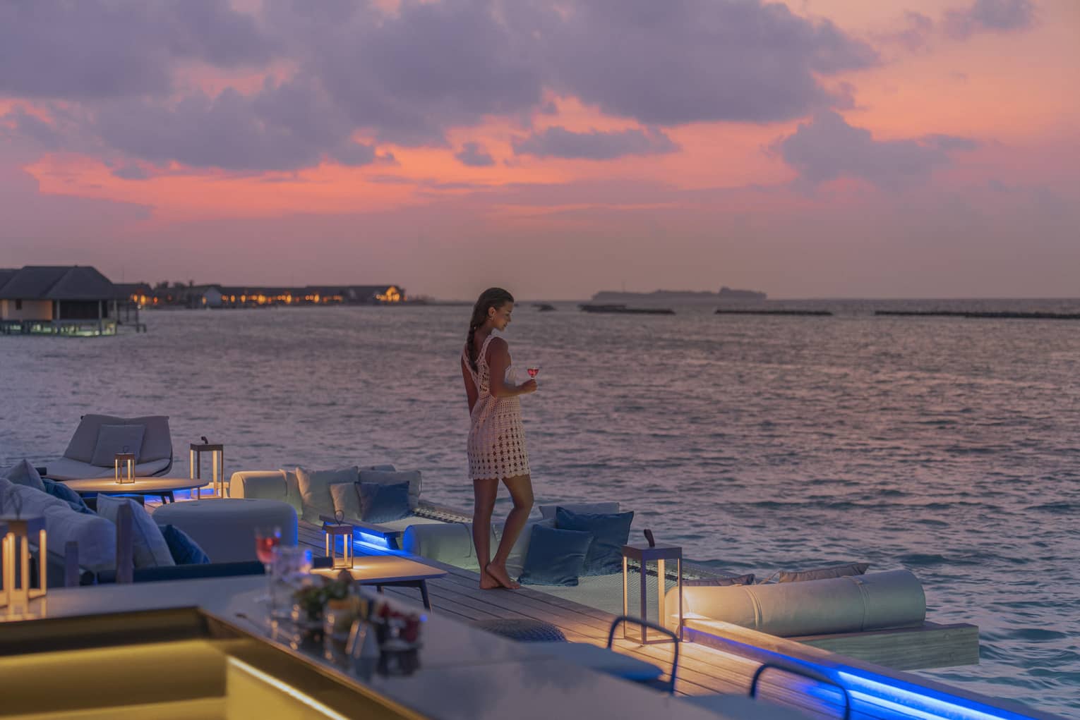 Woman holding cocktail stands at edge of Seabar overlooking the water at sunset