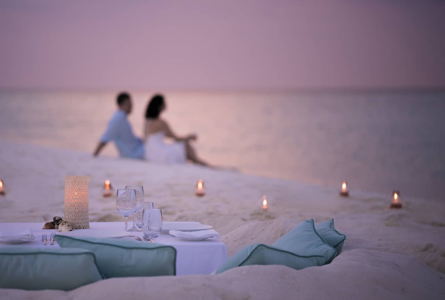 Private dining table in front of sand beach with tealight candles, couple sitting in sand