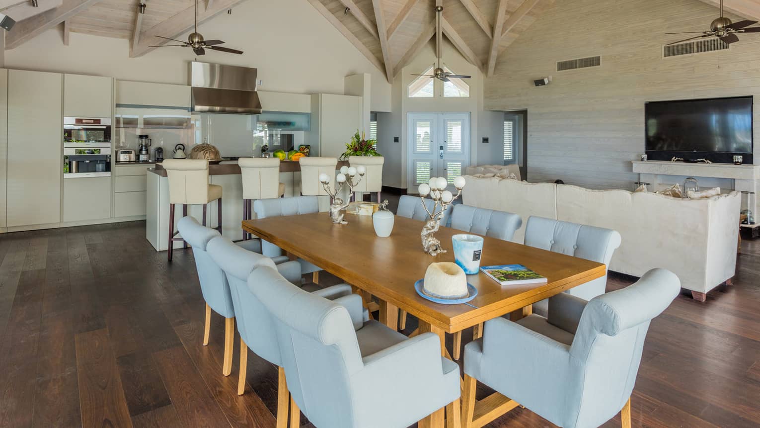 Dining area with wooden table, 8 light-blue upholstered chairs, opening to living area and kitchen