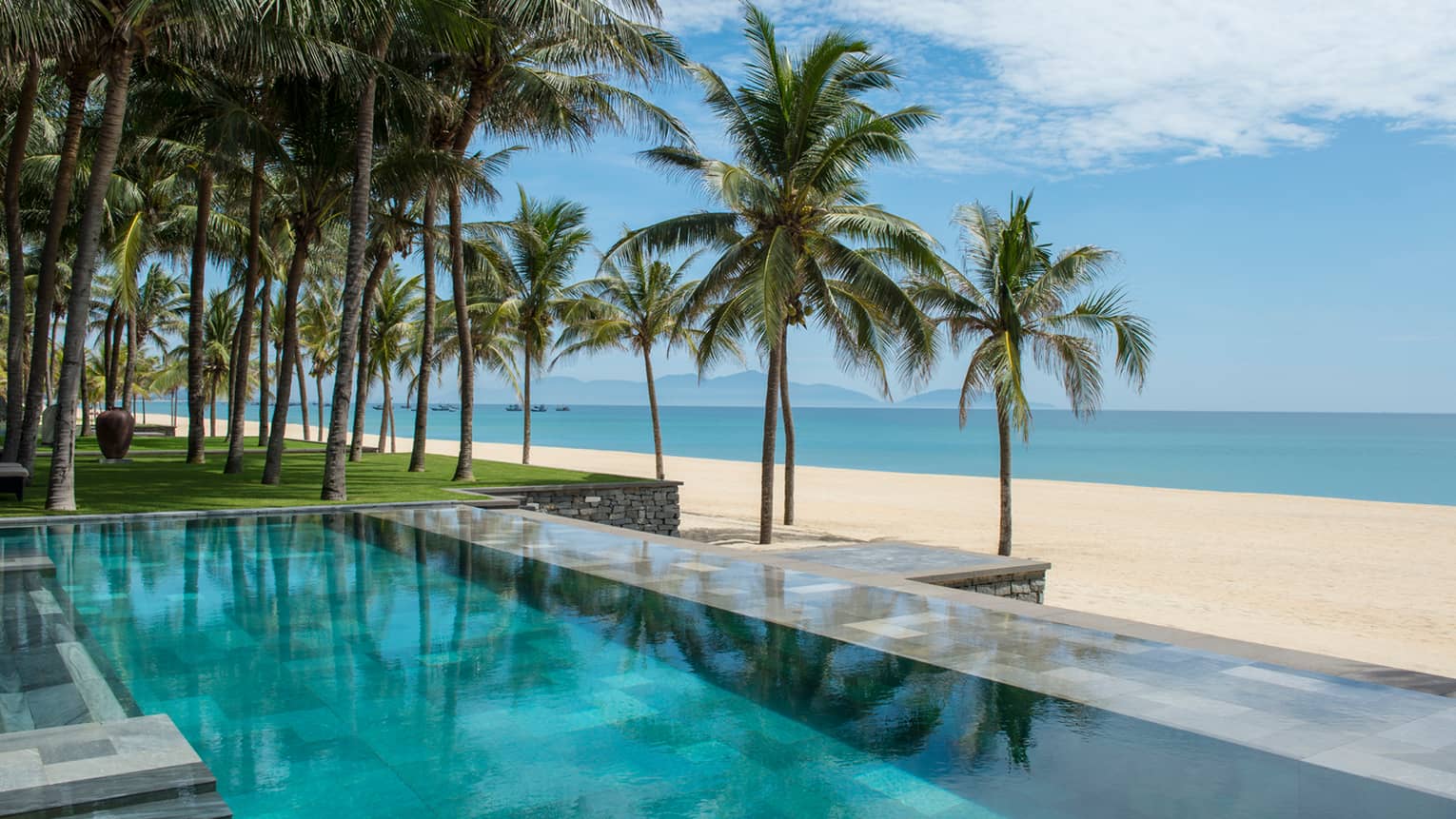 Tall palm trees between stone outdoor swimming pool and white sand beach