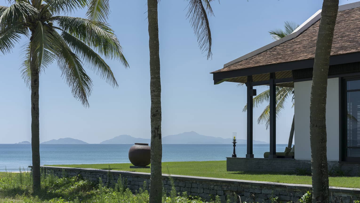 Beachfront villa with palm trees, overlooking a calm ocean and distant islands under a clear blue sky.