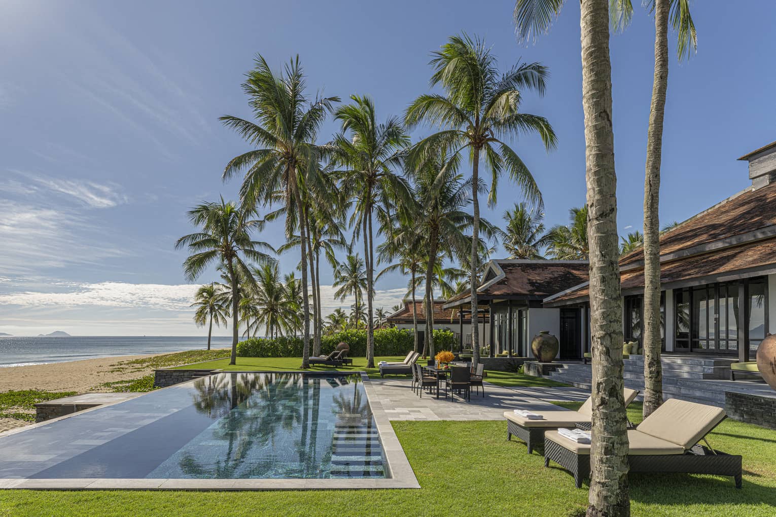 Three Bedroom Beachfront Pool Villa pool area with dining table, lounge chairs, palm trees and blue skies