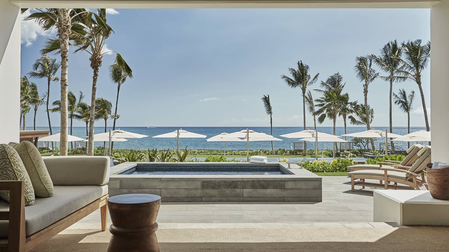 Pacific Suite's luxurious beachfront patio with a comfortable seating area, a private pool and sun loungers facing the ocean, framed by palm trees and umbrellas under a clear sky