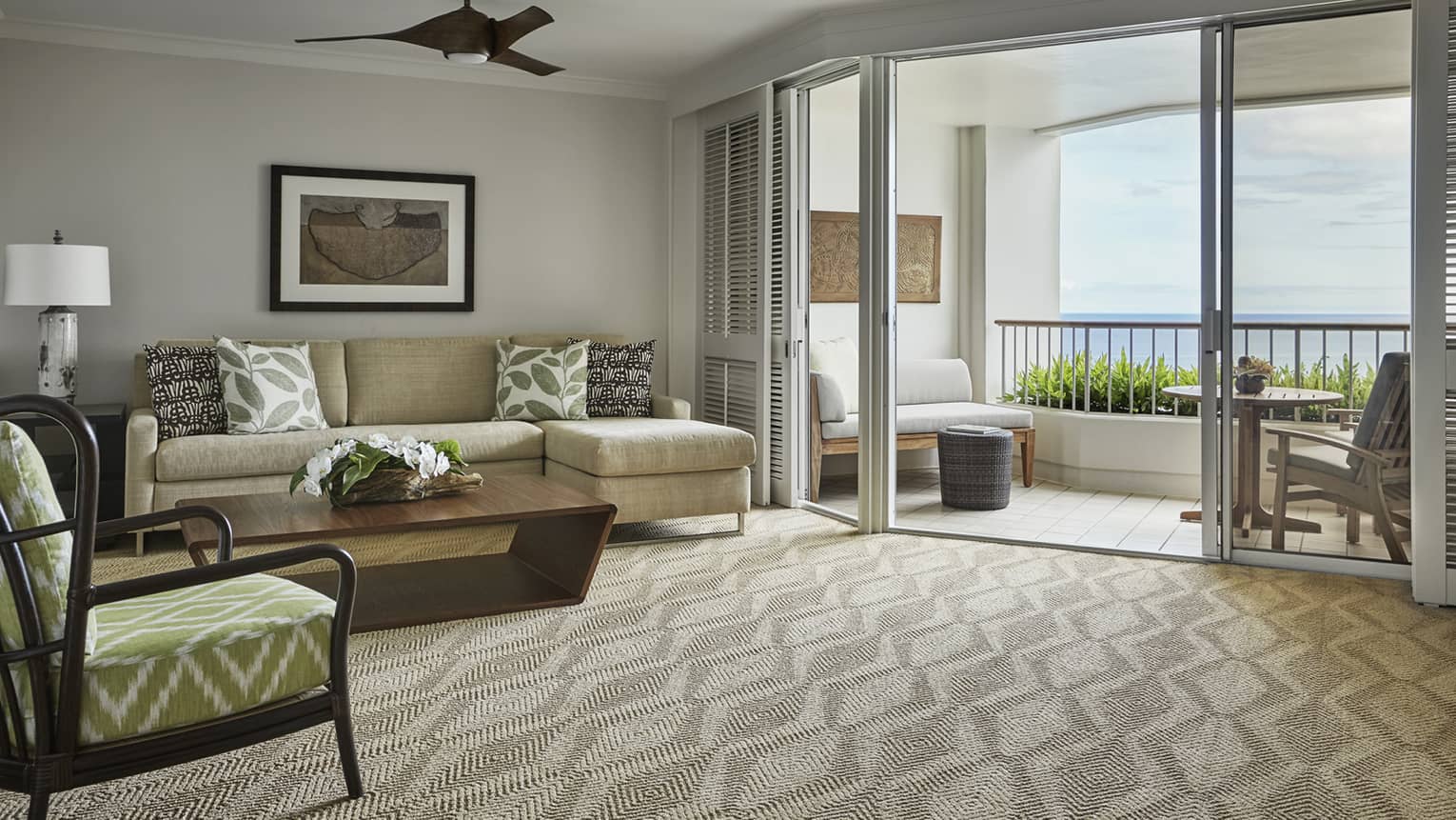 Oceanfront Suite's cosy living room with a beige sectional sofa, patterned cushions and a wooden coffee table. Sliding glass doors open to a balcony with ocean views and comfortable seating.