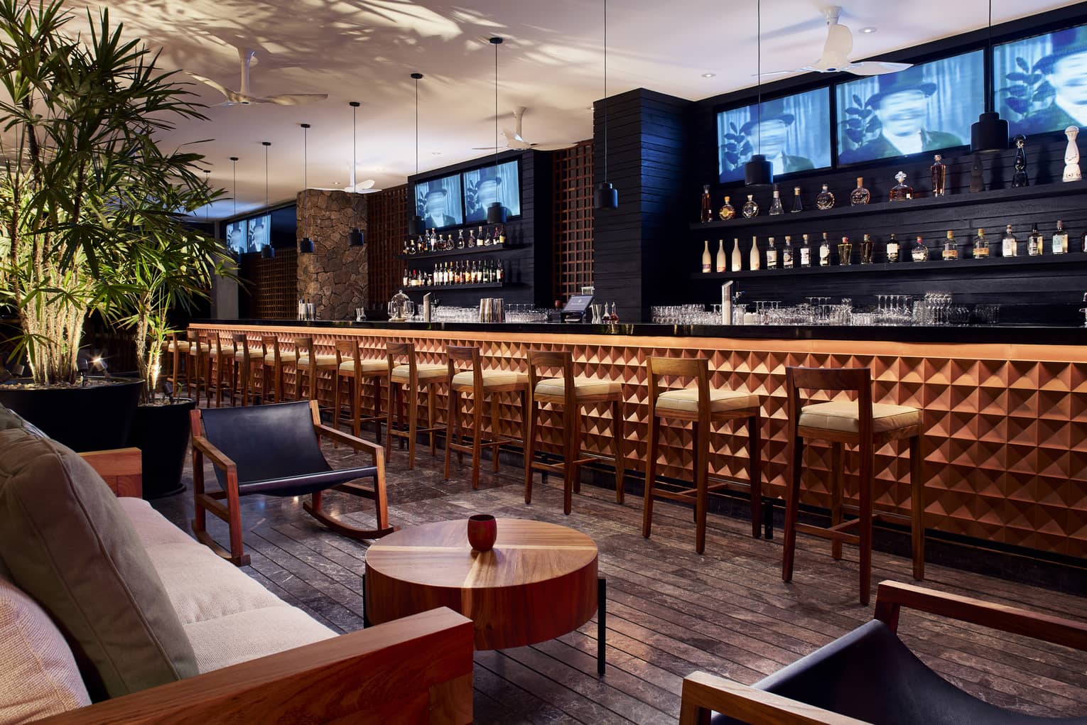 Chairs line a wooden bar with TV screens and shelves of bottles