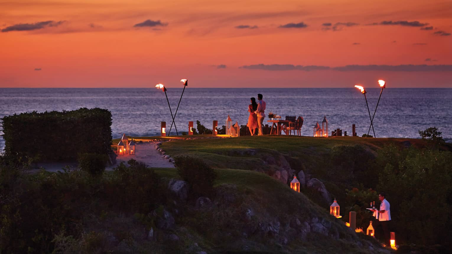 Couple stands on The Rock outpost at dusk, overlooking ocean with private dining table with lanterns, torches 