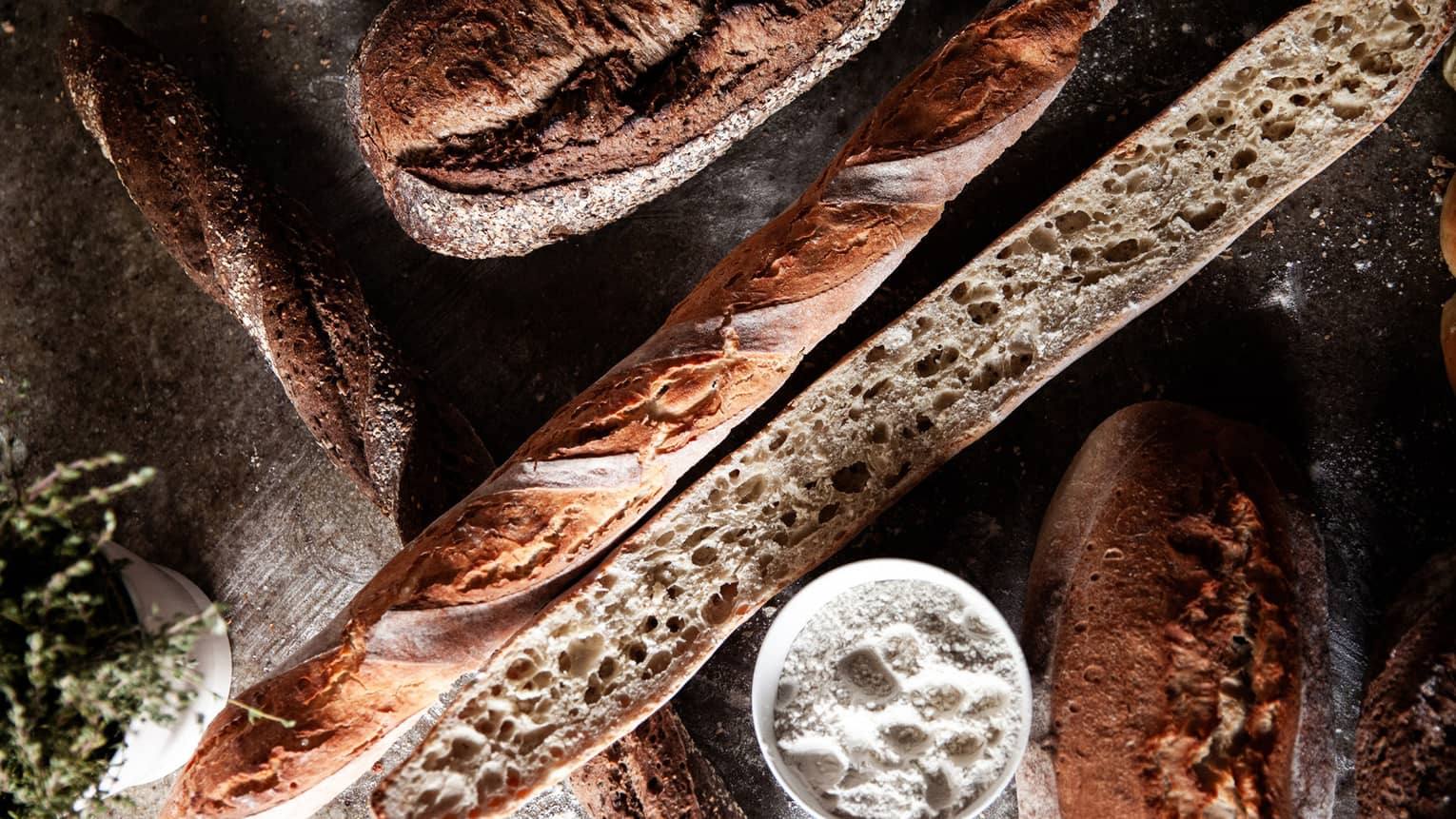 Two baguettes and two loaves of fresh-baked bread sit on a wooden table next to a bowl of salt and a bowl of greens