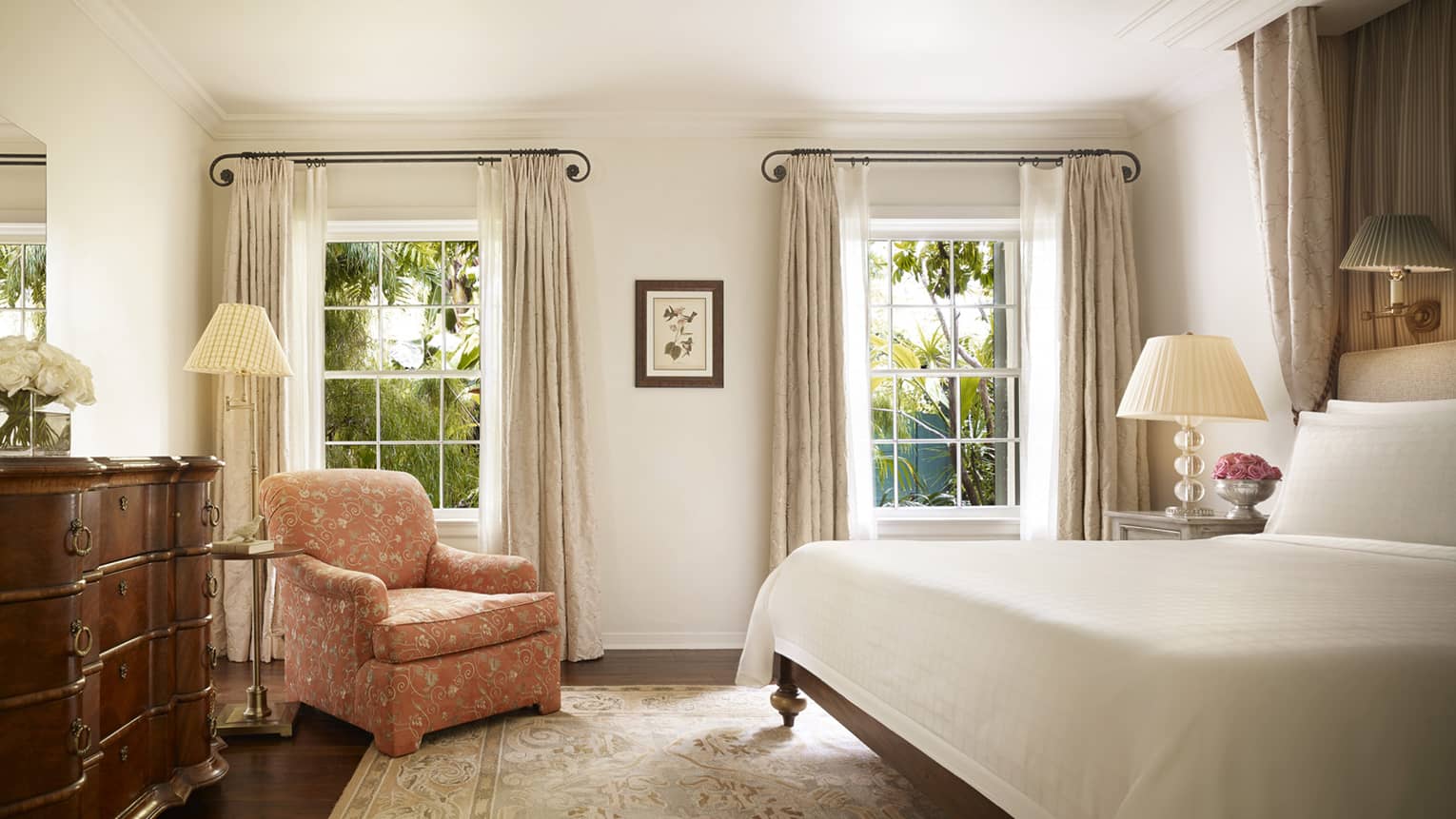 Wood dresser, pink floral armchair by tall sunny windows, nightstand and bed