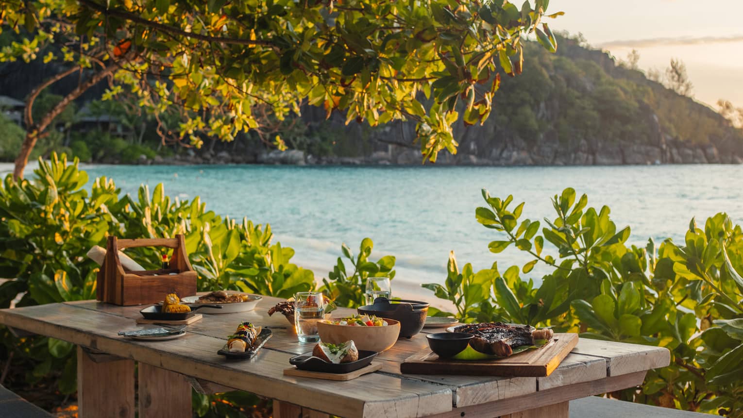 Several dishes on beachside wooden table surrounded by greenery