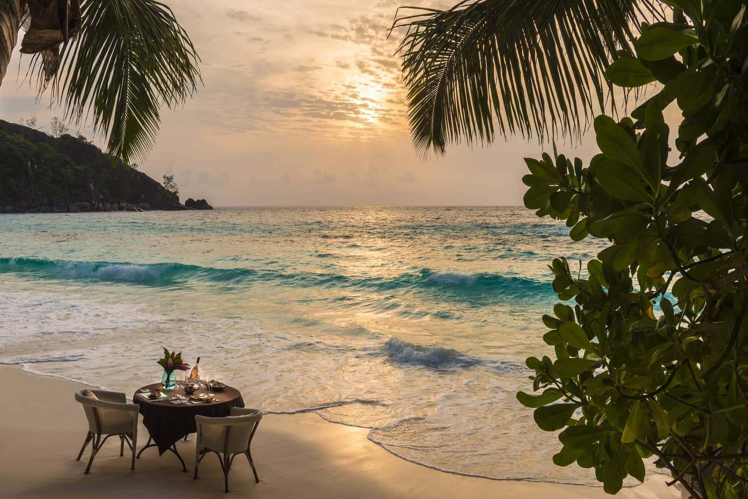 Table for two on sandy beach by ocean's edge at sunset framed by palm trees