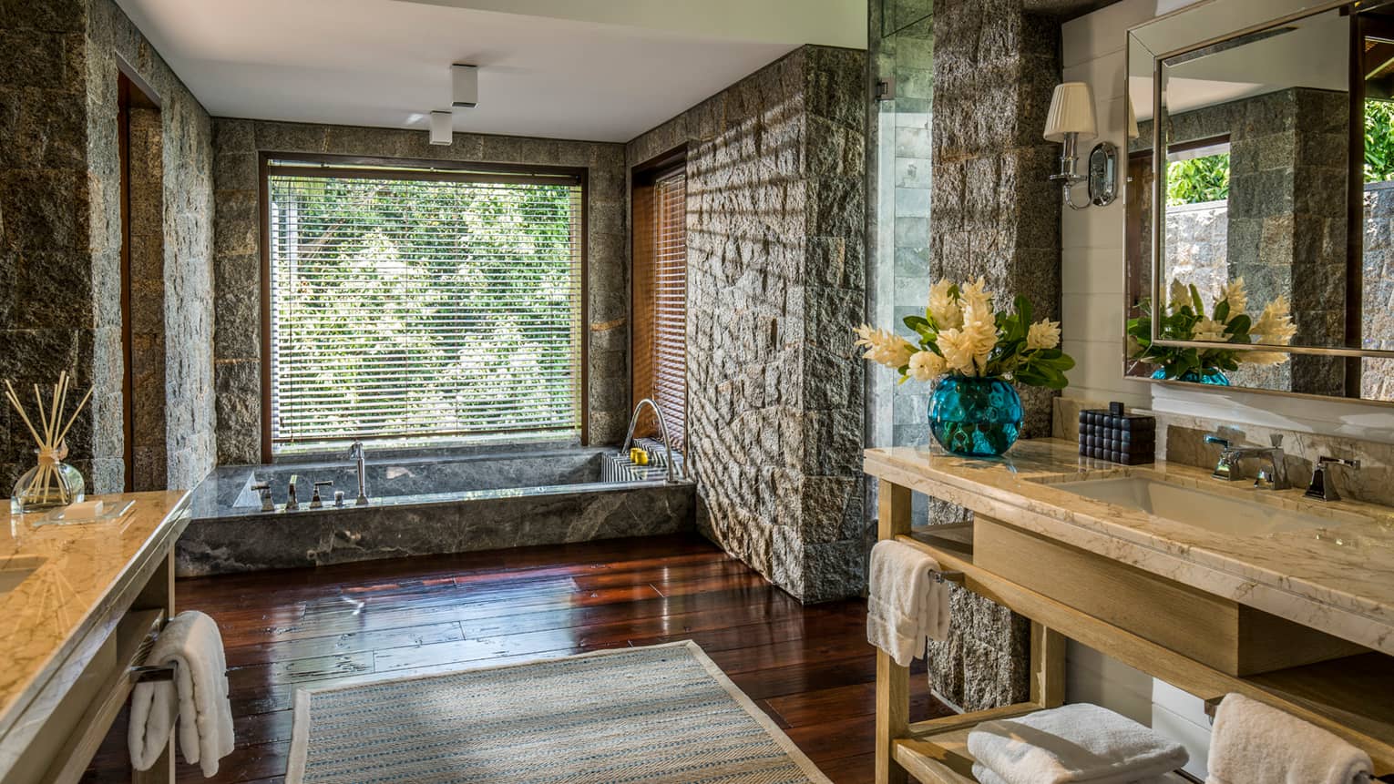 Long vanities in large bathroom with stone walls, sunken marble tub under window