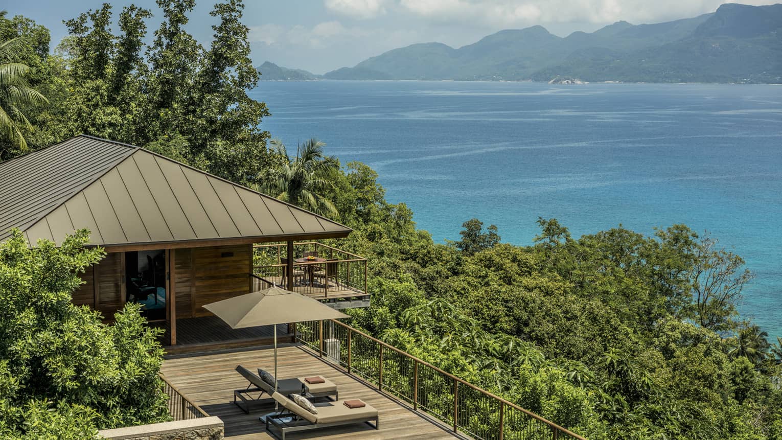 View over wood patio, villa roof on green mountain slope, blue ocean in background