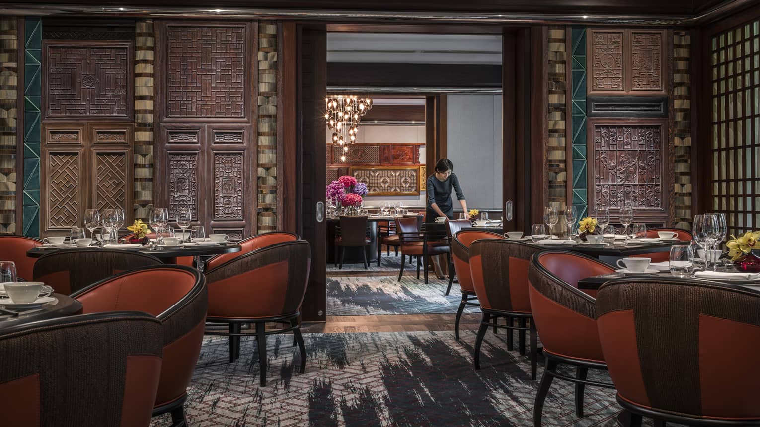 Woman sets table in restaurant with red leather bucket chairs around dining tables, carved wood walls