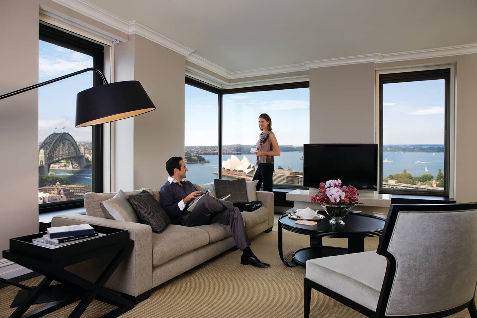 Presidential Suite, woman stands at corner windows, man reads magazine on sofa