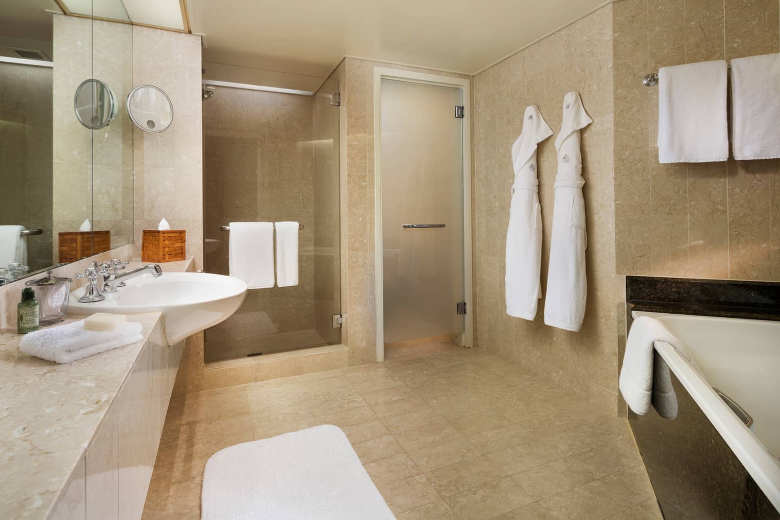Hotel Suite full marble bathroom, two white robes hang by glass walk-in showers, sink