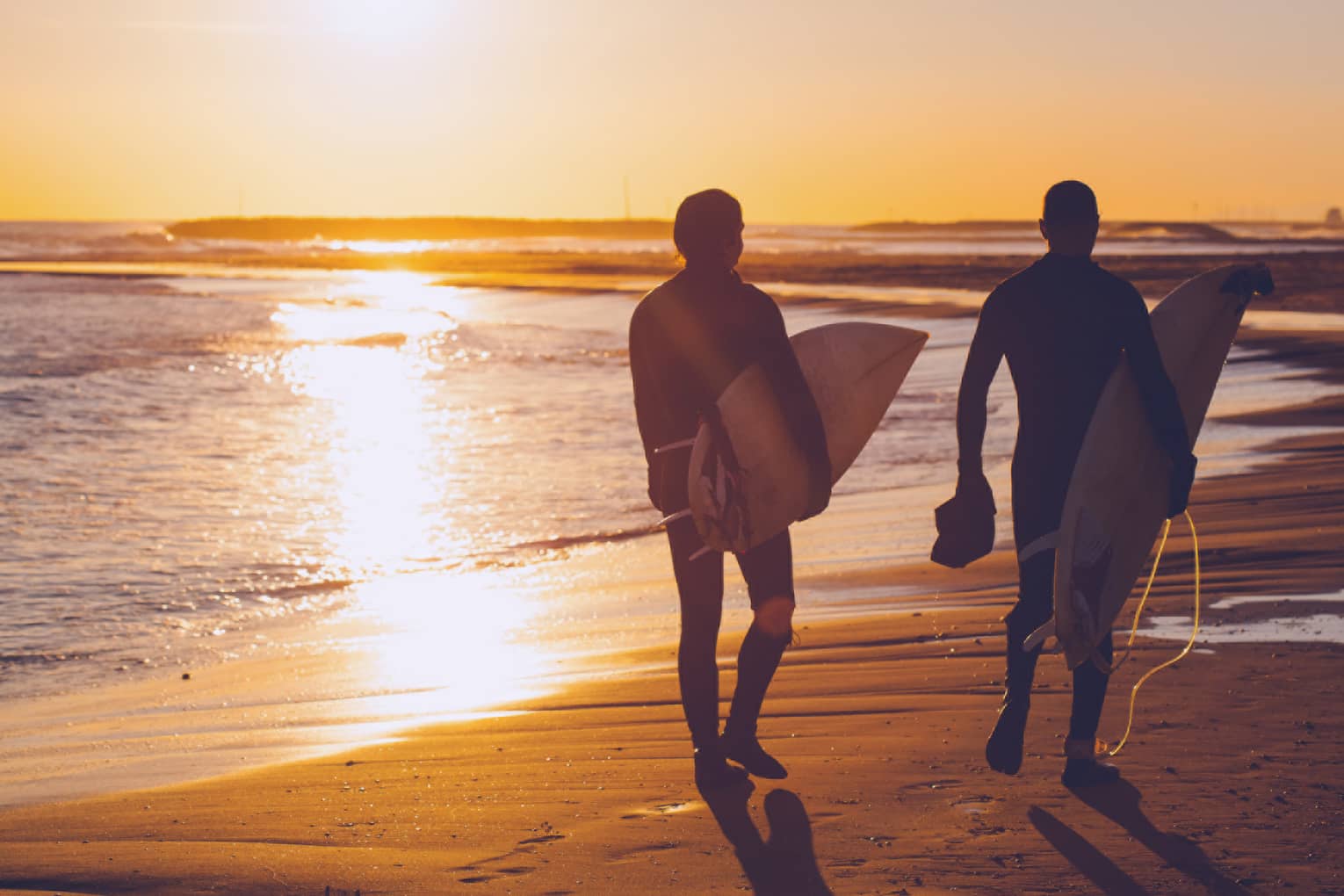 Silhouettes of two people carrying surfboards across beach at sunset