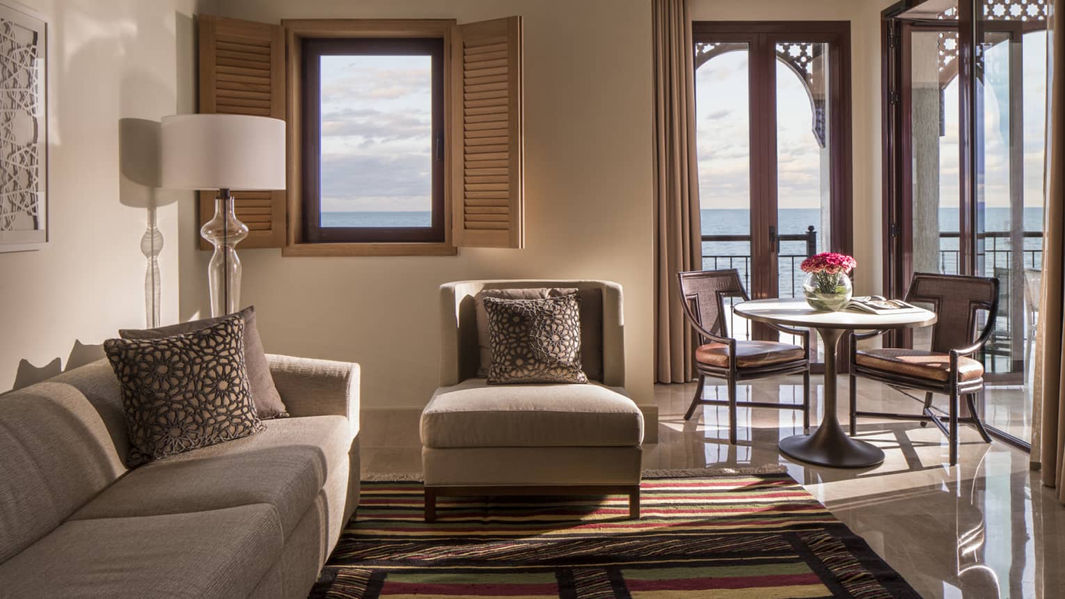 Hotel suite seating area, window with open shutters and doors to corner balcony with view of the sea