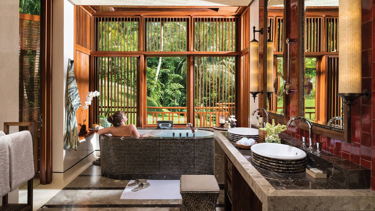 Woman with bare back relaxes in black freestanding spa tub near double sink, wood shutters open to lush garden view