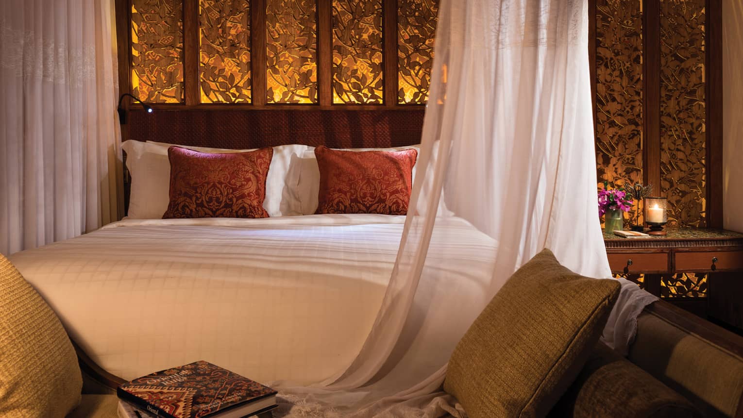Close-up of dimly-lit bedroom, bed with white linens and burgundy pillows, illuminated floral wall behind headboard
