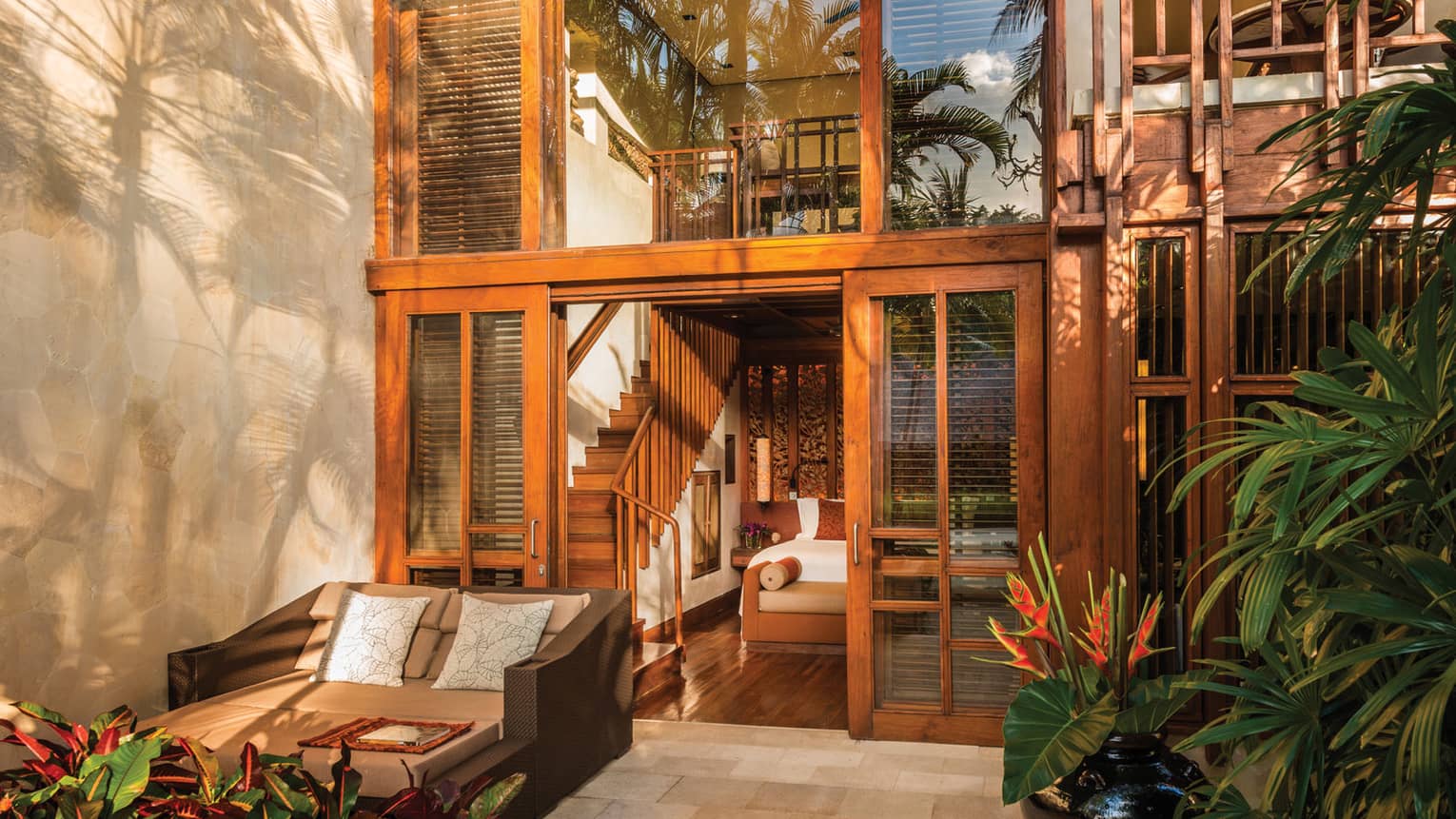 Villa exterior patio looking up at glass windows and doors with wood trim, wood stairwell and bed seen inside