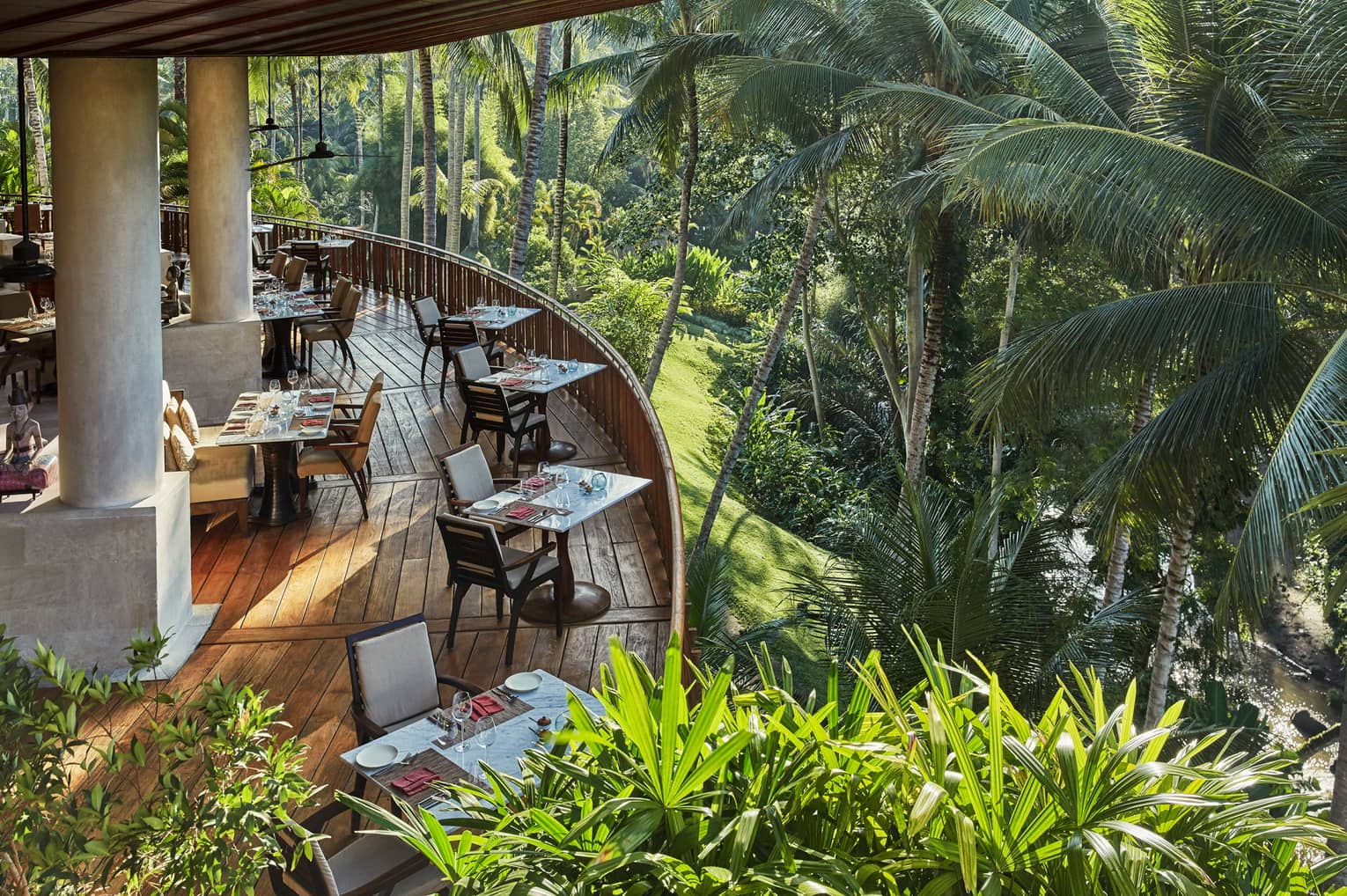 Looking down at curved wood patio of Ayung Terrace outdoor dining room, tables and chairs overlooking palm trees, forest