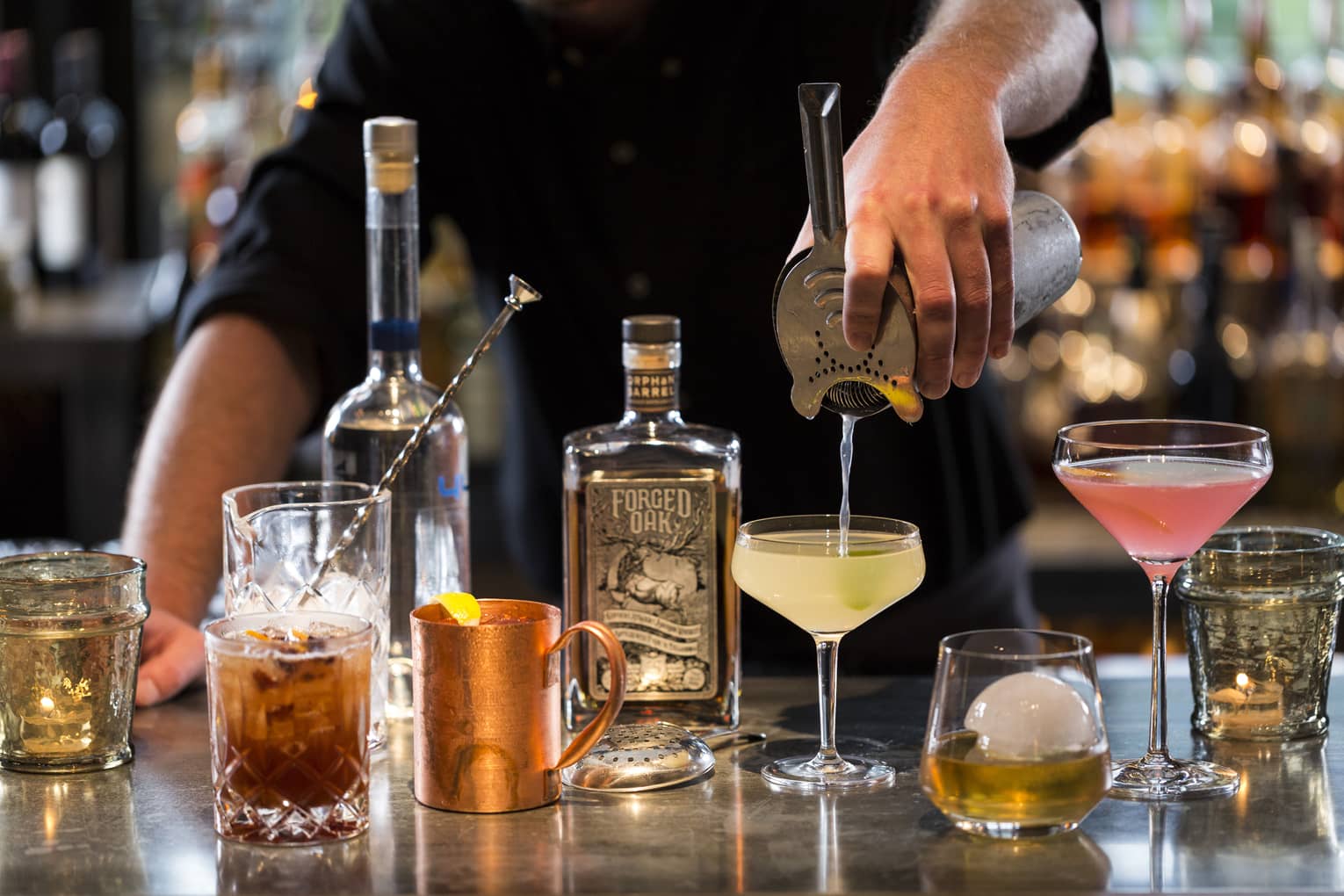 Various cocktail glasses and liquor bottles on bar as mixologist pours martini out of shaker