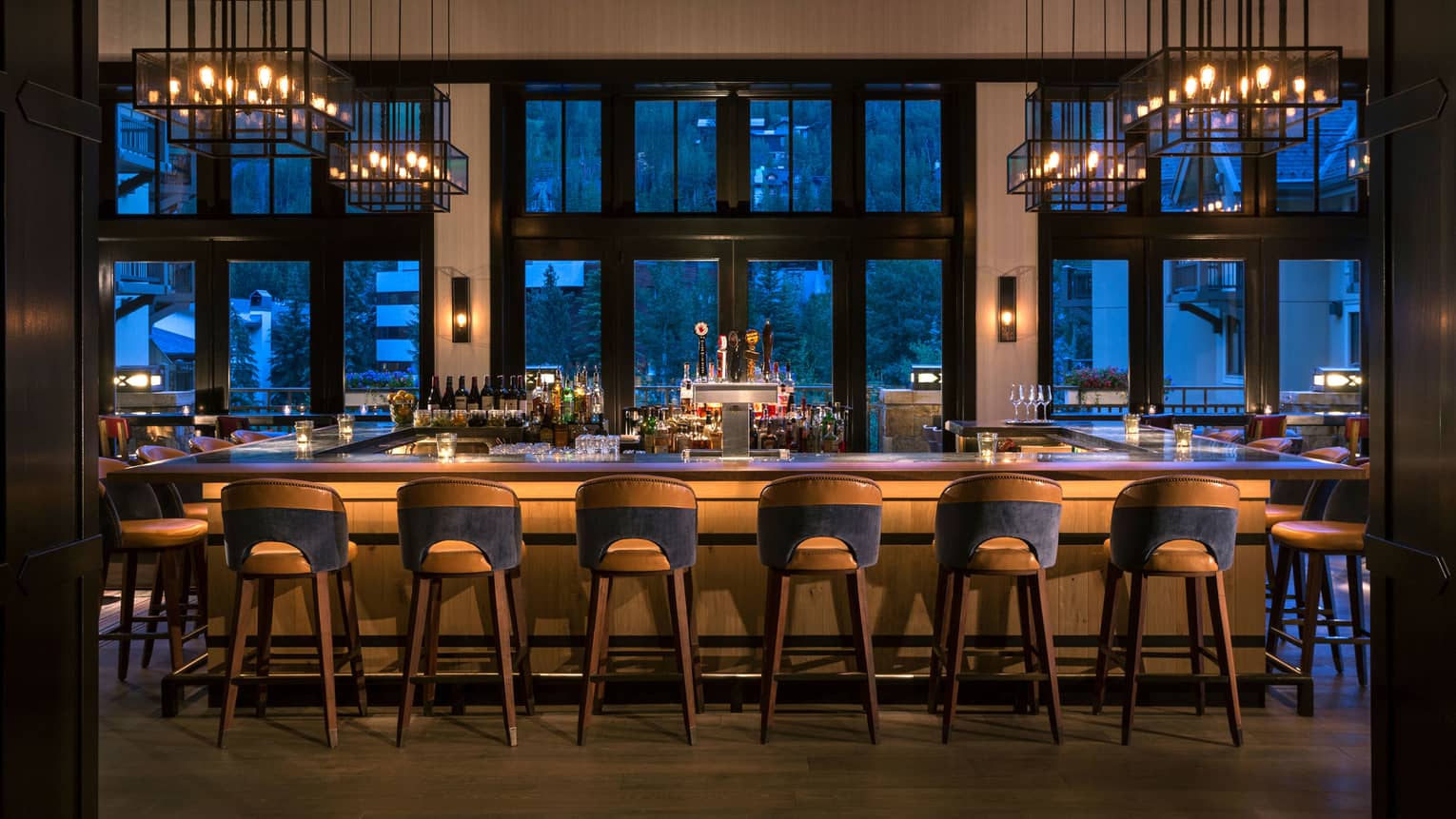 Dimly-lit bar at dusk lined with retro-style wood stools, lantern chandeliers above, large windows