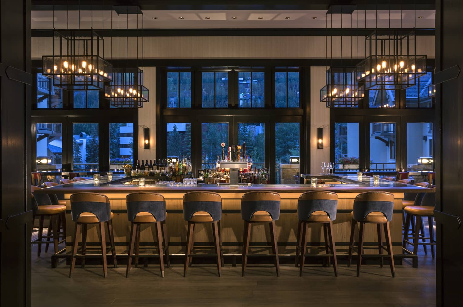 Dimly-lit bar at dusk lined with retro-style wood stools, lantern chandeliers above, large windows