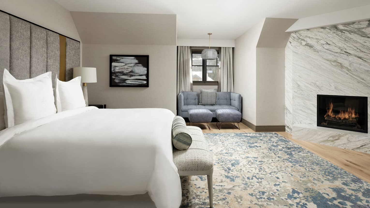 Hotel room bed, small bench and area rug across from marble wall with modern fireplace