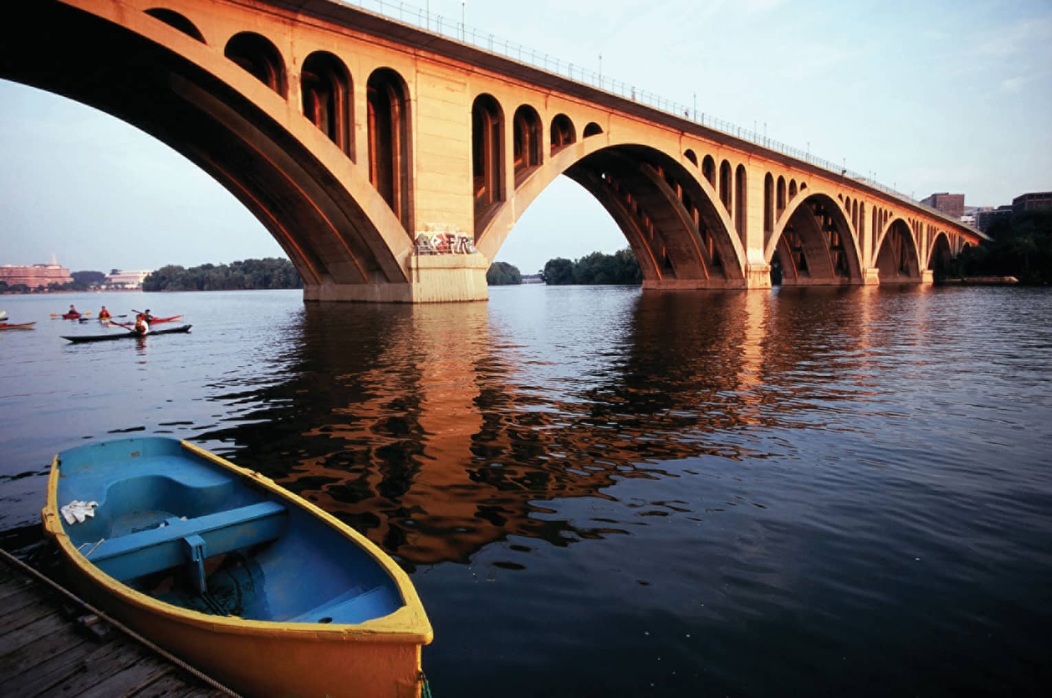 A wide river with a large bridge crossing it, there are people rowing kayaks under the bridge.