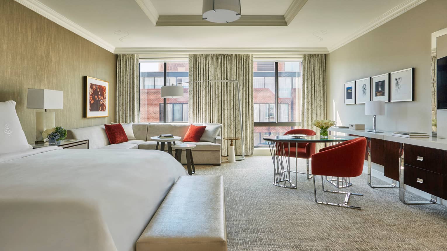 Hotel room corner of bed, bench in front of L-shaped sofa, sunny window, cafe-style dining table with red bucket chairs