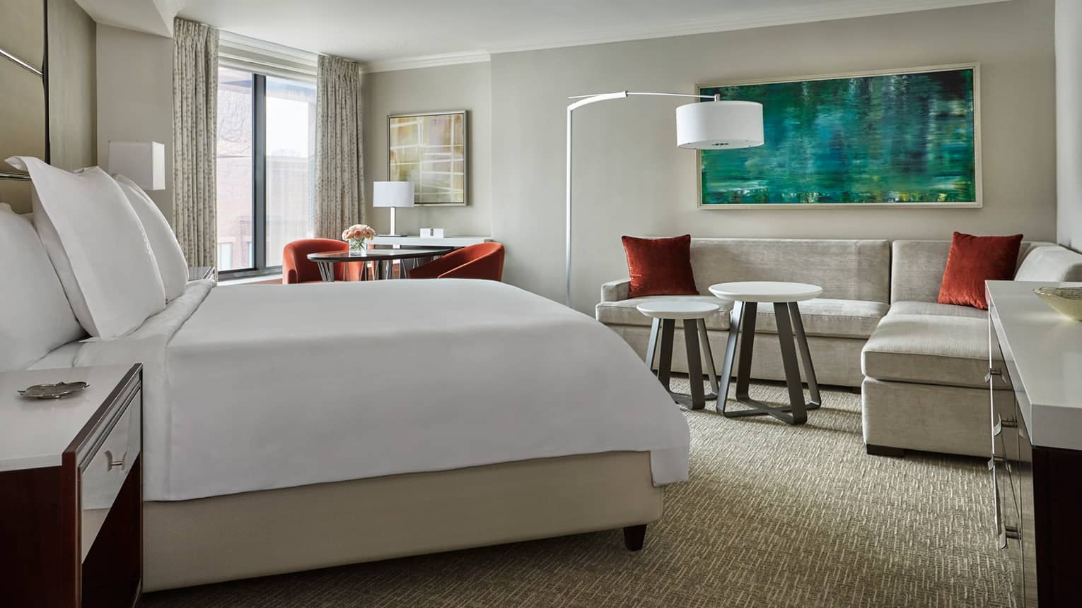 Deluxe Room side of bed with white linens across from L-shaped grey sofa with red pillows, table by window