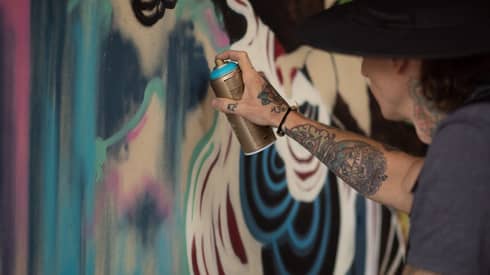 A man with tattoos on his arm and a hat painting a mural with spray paint.