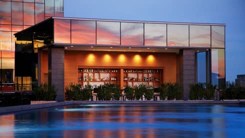 Illuminated swimming pool, wood bar and liquor displays under glass building exterior at sunset