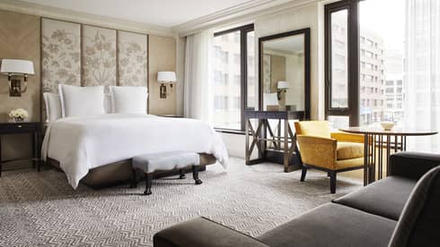 Deluxe City-View room white bed with tall floral fabric headboard, dark wood vanity, yellow armchair and window