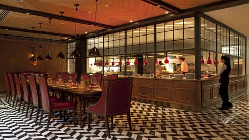 Long 14-person dining table with red leather chairs in front of wood kitchen line, server, black-and-white tile floors