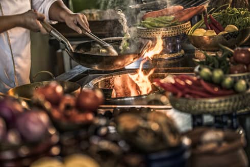 Close-up of chef stirring meals in copper pans over stove, flames 