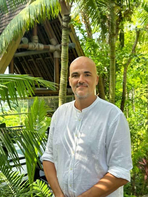 Person in white linen shirt stands outdoors amid palm trees and greenery