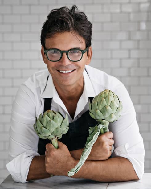 Chef Kenney holding two artichokes in front of a tiled wall.