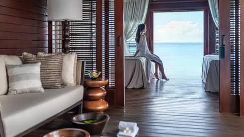 Person sitting on a massage bed in a serene spa treatment room with ocean view, surrounded by cozy seating and wooden décor