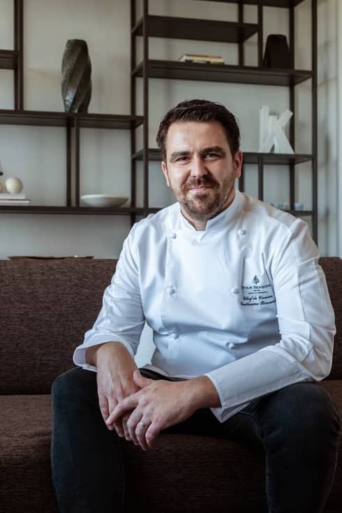 Smiling, bearded man wearing white chef's coat poses for camera while sitting on couch