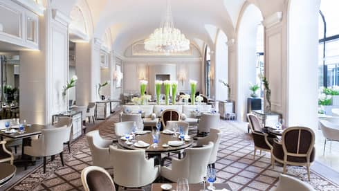 Bright Le George dining room, white chairs around tables under arched pillows, chandeliers