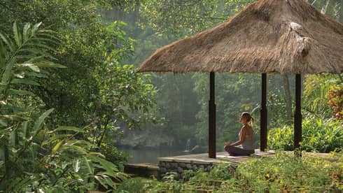 Side view of woman meditating on platform under thatched-roof gazebo, looking out at river and forest