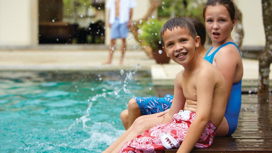 Young boy and girl sit on edge of pool with feet in water