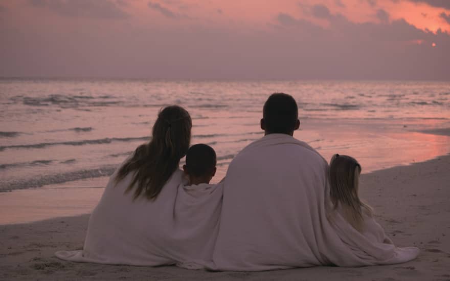 Back of family wrapped in white towels sitting on white sand beach at sunset