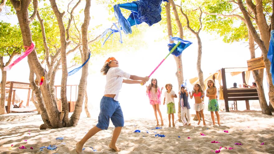 Boy wearing orange blindfold swings stick at blue pinata on beach while five young children cheer him on